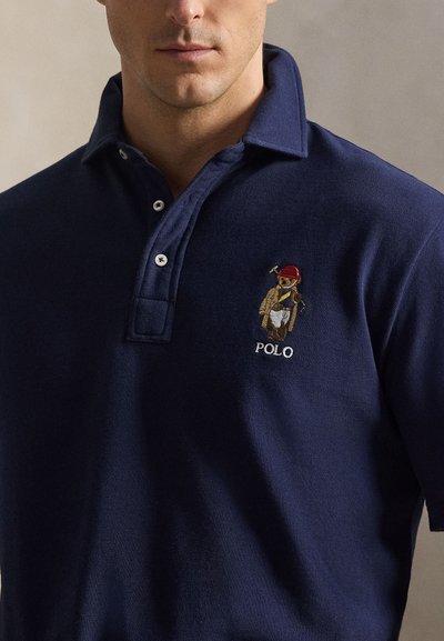 Άνδρας που φοράει μπλε σκούρο πόλο με κεντημένη αρκούδα που κρατάει ρακέτα πόλο στο αριστερό στήθος.