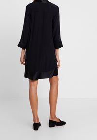 Person som bär en knä-lång svart klänning med satin muddar och fåll, tillsammans med svarta låga klackskor, stående mot en enkel bakgrund.