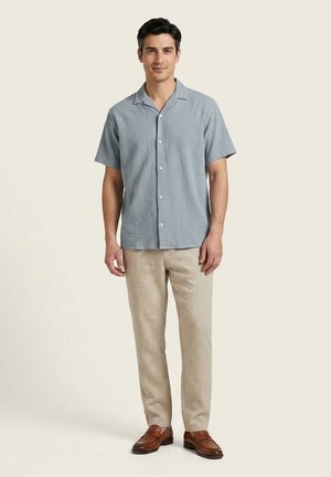 Uomo che indossa una camicia azzurro chiaro a maniche corte con bottoni, pantaloni beige e mocassini in pelle marrone, in piedi davanti a uno sfondo semplice.