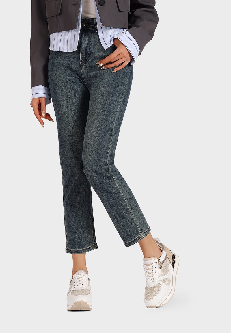 Giacca grigia leggera e corta sopra una camicia bianca a righe blu, abbinata a jeans dritti in denim scuro e sneaker beige.