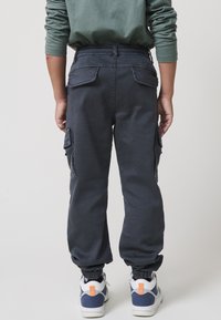 Mornarski cargo hlače z elastičnim pasom in manšetami, s stranskimi in zadnjimi žepi, v kombinaciji z belimi supergami in oranžnimi akcenti.