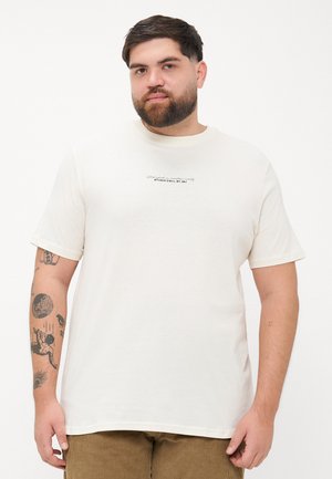 Mand med skæg iført en hvid t-shirt med lille tekst og brune bukser, der viser flere tatoveringer på sin venstre arm mod en hvid baggrund.