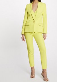 Vrouw in een heldergele, op maat gemaakte blazer en bijpassende slim-fit broek met gouden strappy hoge hakken.