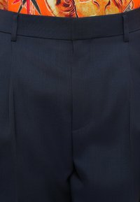 Tummansinisiä housuja, jotka on valmistettu tekstuurikankaasta. Ne ovat litteällä edellä, yhdessä laskoksella ja vyölenkeillä, ja niissä on hieman räätälöity istuvuus.