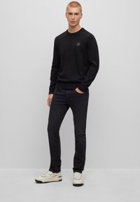 Felpa nera girocollo con una piccola toppa logo, abbinata a jeans neri slim-fit e sneakers bianche. Texture liscia, design minimalista.