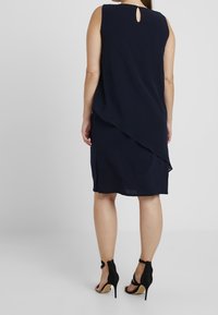 Marinblå ärmlös klänning med draperad skärning, nyckelhållsdetalj på ryggen, mjukt tyg och sidslitsar, i kombination med svarta snörade högklackade skor.