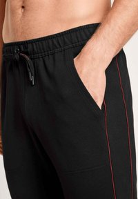 Pantalons de sport noirs avec une taille à cordon de serrage, présentant des poches latérales et des rayures rouges le long des coutures. Matière en mélange de coton, texture lisse.
