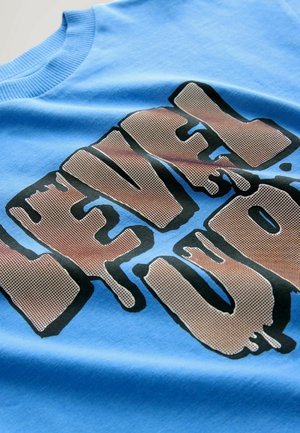 Tricou din bumbac albastru, cu un text grafic mare, îndrăzneț, în negru și marron, care spune "LEVEL UP", având un efect de picurare și o textură netedă.
