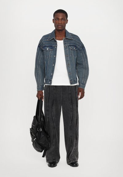 Denimjakke med lodrette striber, to brystlommer, båret over en hvid skjorte. Løse, sorte bukser med dekorativ syning, holdt af en person med en sort taske.