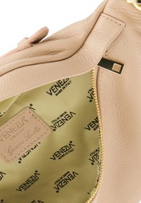 Indersiden af en håndtaske i cremefarvet læder med guld lynlås og beige foring, der præsenterer gentagne "VENEZIA"-mærkater. Etiket med teksten "Ægte læder."