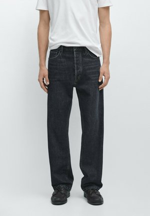 Mann trägt lockere schwarze Jeans aus Denim, schwarze Schuhe und ein schlichtes weißes T-Shirt, steht vor einem neutralen Hintergrund.