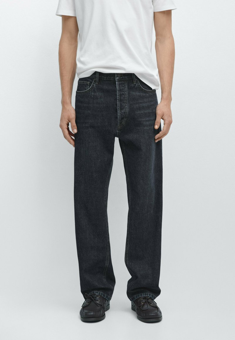 Mand iført løse sorte denimjeans, sorte sko og en enkel hvid t-shirt, stående foran en neutral baggrund.