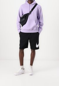 Violetti huppari mustalla olkalaukulla, mustat shortsit, joissa suuri valkoinen Nike-logo, ja valkoiset urheilukengät. Rentoutunut urheiluasu.