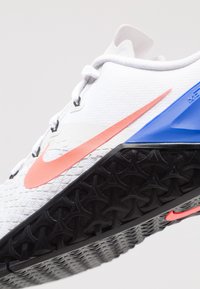 Zapatilla deportiva blanca con una parte superior texturizada, un logo de Nike en rosa, detalles en azul en el talón y una suela de tracción negra.
