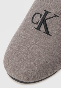 Papuci gri din fetru cu logo-ul brodat „CK” în negru. Design cu vârf rotund și talpă din cauciuc negru pentru aderență. Textură moale și detalii minime.
