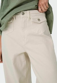 Pantalons beige clair à coupe ample, avec des poches avant et arrière, fermeture à bouton argenté et tissu texturé. Associés à un haut vert.