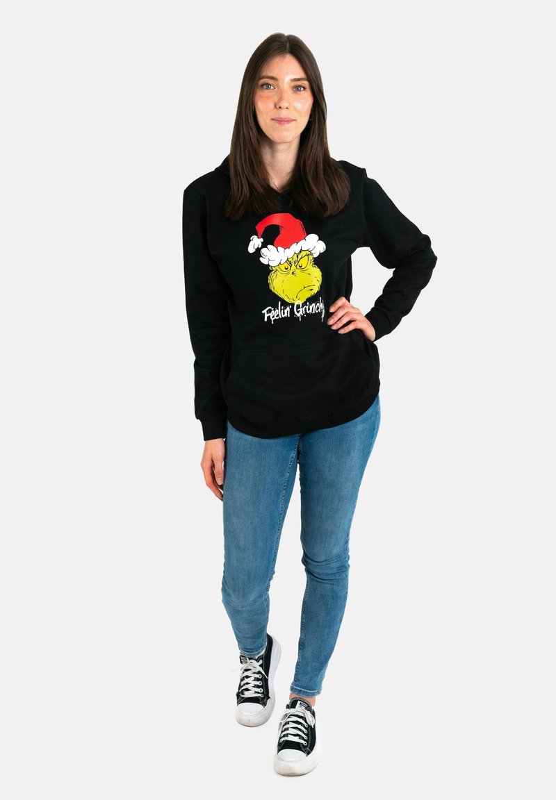 Black sweatshirt featuring a graphic of the Grinch in a red Santa hat with "Feelin Grinchy" text, paired with light blue jeans and black sneakers.