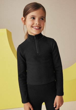 Haut noir à manches longues et côtelé avec un col haut et une fermeture éclair demi-longueur, associé à un legging côtelé assorti. Design minimaliste sur un fond doux.
