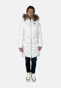 Cappotto bianco imbottito con cappuccio in pelliccia sintetica, design trapuntato, zip frontale, due tasche laterali, indossato sopra jeans scuri e sneakers.