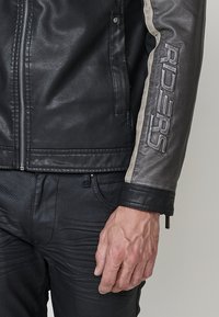 Crna kožna jakna sa zipom sprijeda, teksturiranim akcentima i krem trakama sa strane na kojima je urezano "RIDERS". Model nosi crne hlače.