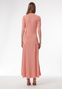 Lang, geplooid jurk in lichtroze met driekwart mouwen en een aansluitende lijfje. De achterkant heeft een vaste bovenkant die overgaat in geplooide stof.