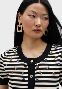 Crno-bijeli prugasti kardigan s okruglim izrezom, ukrašen šarenim draguljima i zlatnim perlicama, s prednjim kopčanjem na gumb.