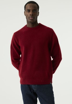 Man standing against plain background, wearing a red crew neck sweater and blue jeans, hands behind his back, facing forward.