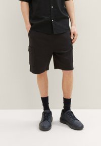 Muž v černých cargo šortkách, černých ponožkách, černých teniskách a černé košili s krátkým rukávem stojí proti jednolitému pozadí.