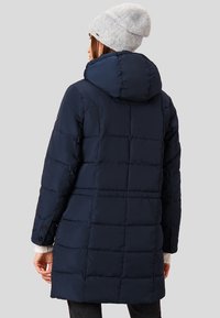 Námořnická modrá prošívaná bunda s kapucí, s prošívaným designem a knoflíky na rukávech. Je na sobě světle šedá pletená čepice.