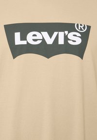 T-shirt en coton beige arborant un grand logo Levi's vert foncé avec des lettres blanches et un design incurvé emblématique.