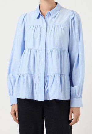 Mujer que lleva una blusa de capas azul claro con mangas largas abullonadas y cuello, combinada con pantalones negros.
