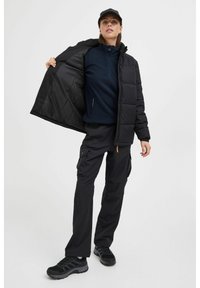 Svart isolert jakke med glidelås, paret med en marineblå genser og svarte cargo-bukser. Modell med svarte sko og caps.