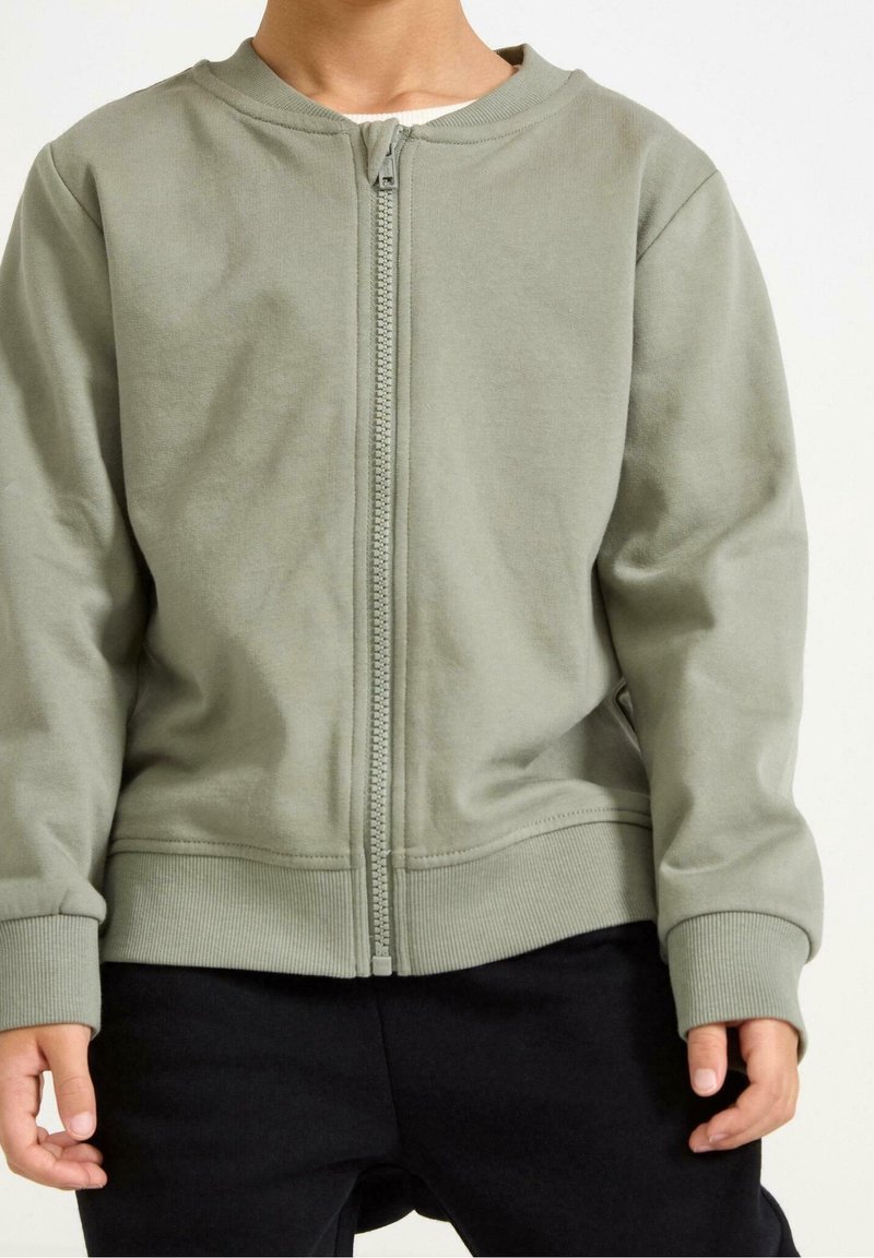 Personne portant une veste zippée vert clair avec des poignets et un ourlet côtelés, associée à un pantalon noir, debout devant un fond uni.