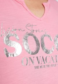 Rosa kortärmad T-shirt med metalliskt silverfärgad text som säger "Lisbon Stories SOCC ON VACATION" och en V-ringning.