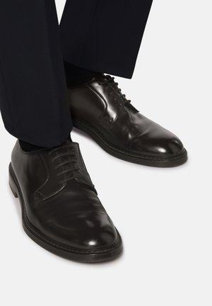 Zapatos de vestir de cuero negro con una superficie pulida, puntera redondeada y diseño clásico de cordones, combinados con pantalones oscuros.