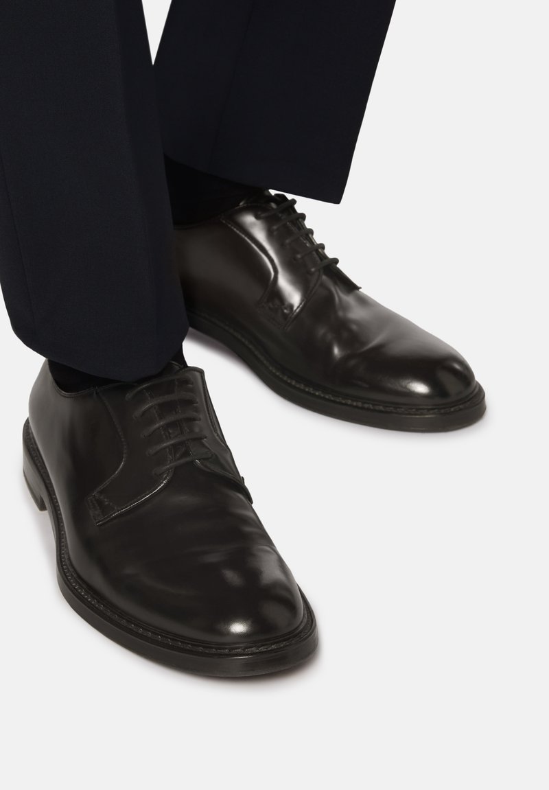 Scarpe eleganti in pelle nera con superficie lucida, punta arrotondata e design classico con allacciatura, abbinate a pantaloni scuri.