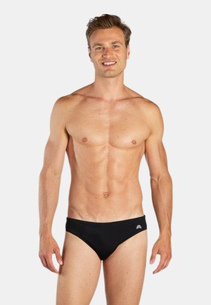 Uomo sorridente in piedi con slip da nuoto neri contro uno sfondo bianco, in posa con le braccia rilassate lungo i fianchi.