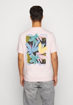Mand iført lyserød T-shirt med farverigt abstrakt bladdesign og tekst med ordene ""Born Free, Stay Free, Made with Love"" på bagsiden.