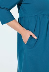 Turkusowa sukienka wykonana z miękkiego materiału, z marszczoną talią i bocznymi kieszeniami. Niezwykłymi detalami są rękawy o długości 3/4 oraz gładka faktura.