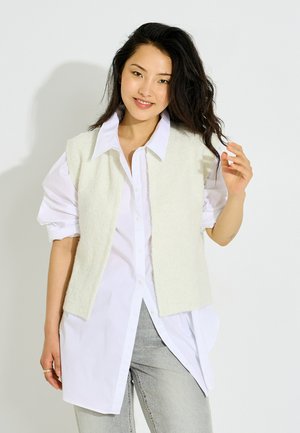 Joven mujer con cabello oscuro lleva una camisa blanca con botones, un suéter sin mangas color crema y pantalones gris claro, sonriendo mientras se ajusta el cabello.