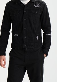 Zwarte denimjas met twee borstzakken, knoopsluiting en geborduurde details. Het ontwerp bevat een witte logo-accent en een geweven afwerking.