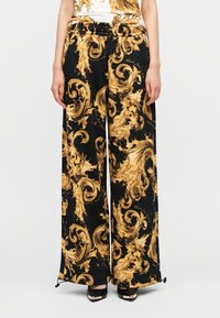 Μαύρο παντελόνι με φαρδιά παντελόνα και χρυστό στροβιλιζόμενο μπαρόκ σχέδιο, με ελαστική μέση, σε συνδυασμό με μαύρες μυτερές γόβες και μερικώς ορατό τοπ με σχέδιο.