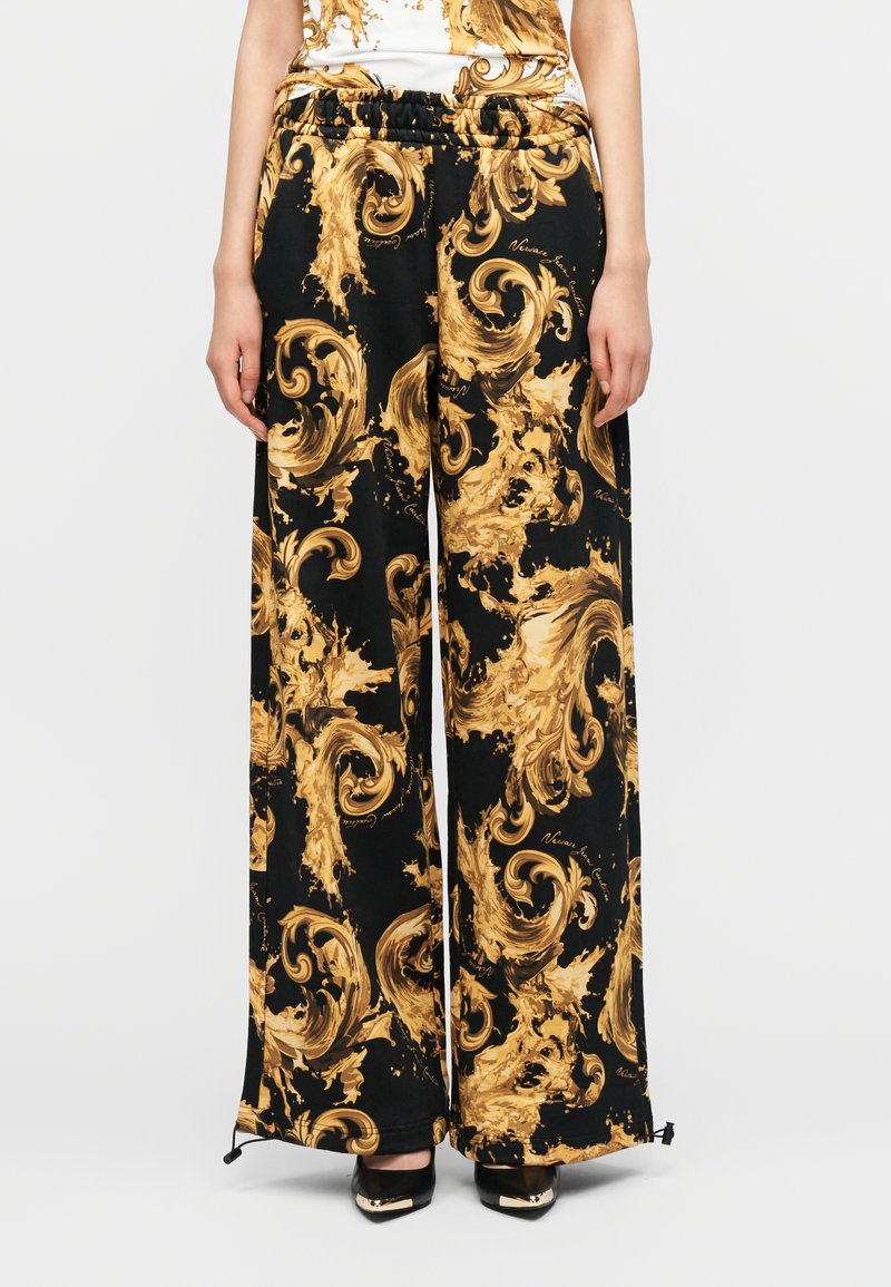 Μαύρο παντελόνι με φαρδιά παντελόνα και χρυστό στροβιλιζόμενο μπαρόκ σχέδιο, με ελαστική μέση, σε συνδυασμό με μαύρες μυτερές γόβες και μερικώς ορατό τοπ με σχέδιο.