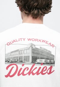 Λευκό γραφικό t-shirt με μια vintage ασπρόμαυρη εικόνα ενός εργοστασίου, με κόκκινο κείμενο που γράφει "QUALITY WORKWEAR" και "Dickies."