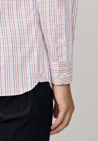 Chemise à carreaux multicolore avec des lignes rouges et bleues sur fond blanc, dotée de poignets boutonnés et d'un ourlet incurvé. Jeans en denim visibles.