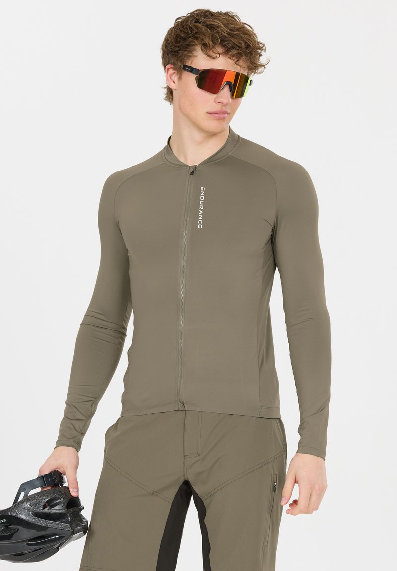 Junger Mann in olivgrünem Fahrrad-Outfit und rot getönten Sportsonnenbrillen, der einen schwarzen Fahrradhelm in einer Hand hält.