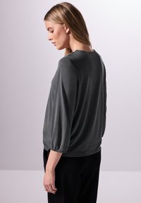 Graue Bluse mit weichem, leicht glänzendem Material, ausgestattet mit drei Viertel Puffärmeln und einem runden Ausschnitt. Sanfte, lockere Passform.