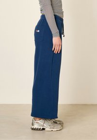 Pantalon de survêtement bleu, à jambes larges, avec taille à cordon et poche latérale. Tissu texturé et accents colorés brodés sur la poche.