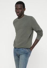 Olivinvihreä ribbikuvioinen neulepaita, jossa on pyöreä pääntie ja pitkät hihat, yhdistettynä vaaleansinisiin farkkuihin.