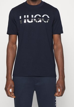Uomo che indossa una maglietta blu navy scuro con la scritta riflettente argento "HUGO" sul petto, abbinata a pantaloni blu navy coordinati.